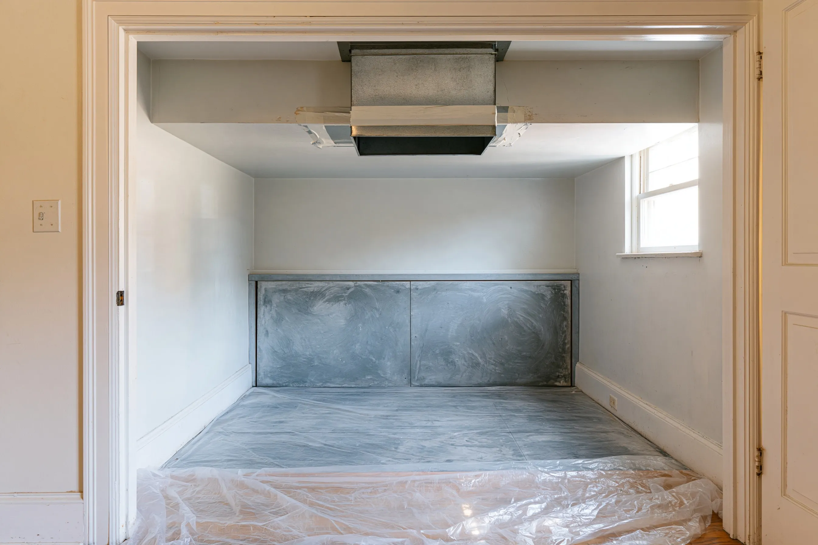 An empty utility closet with a metal duct at the ceiling, a gray panel on the back wall and plastic sheeting on the floor